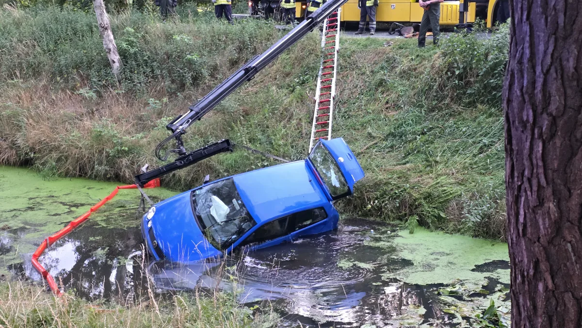 Ein blauer Pkw liegt zur Hälfte in einem Graben und wird mit einem Kran herausgezogen. Feuerwehrkräfte stehen am Rand und beobachten die Bergung.