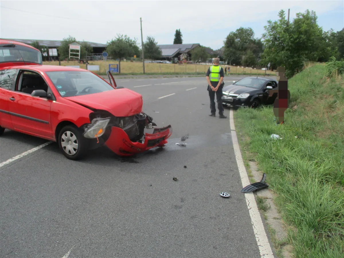 Zwei Autos stehen nach einem Zusammenstoß auf einer Landstraße, wobei ein rotes Auto am stärksten beschädigt ist. Ein Polizist steht daneben.