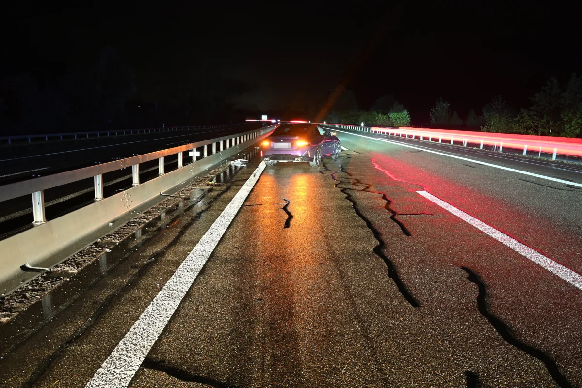 Ein Auto, das bei Nacht auf einer nassen Autobahn steht, zeigt Beschädigungen am Heck. Die rechte Fahrbahnbegrenzung ist deutlich sichtbar.