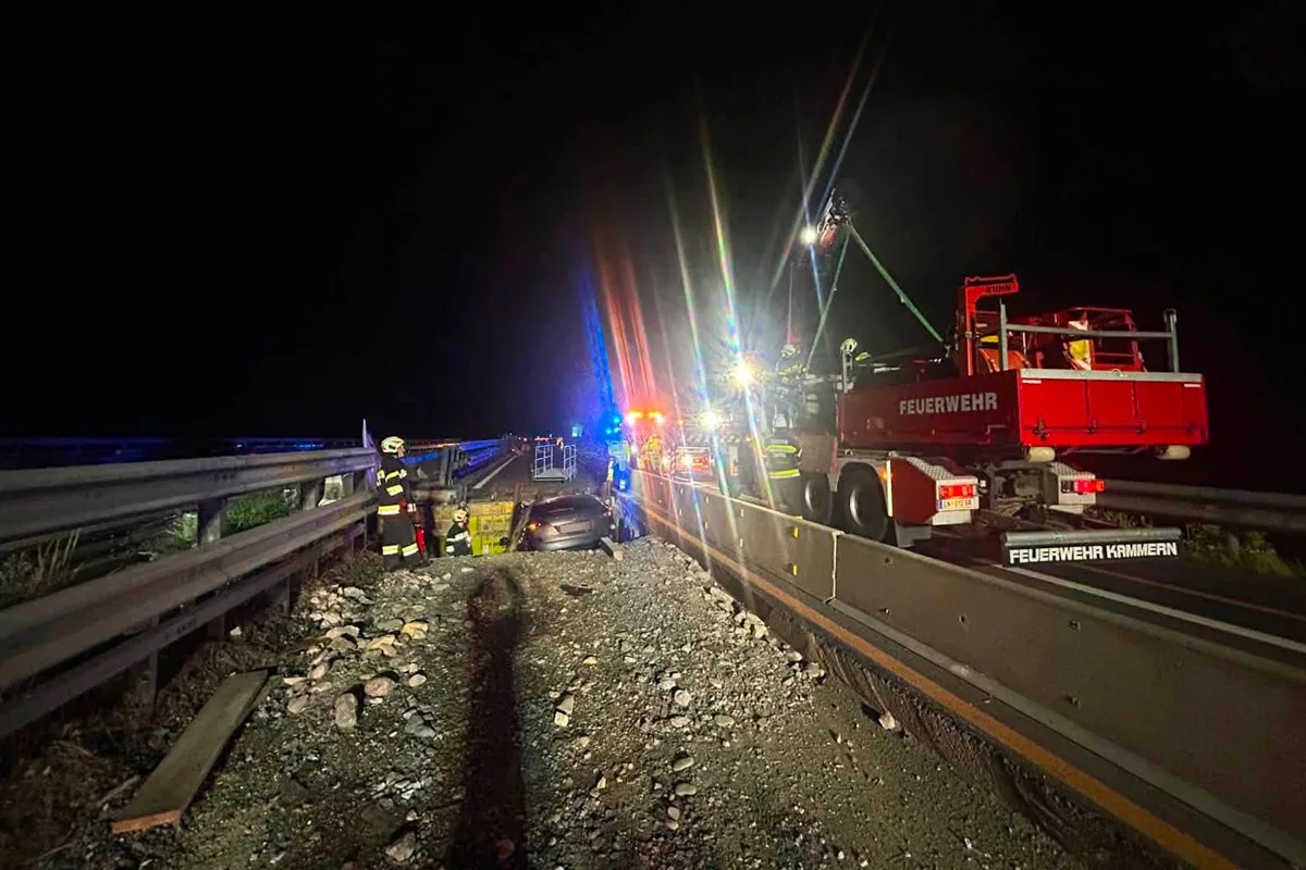 Mehrere Feuerwehrleute sind nachts auf einer Straße im Einsatz. Ein Fahrzeug steht neben der Leitplanke, beleuchtet von Einsatzfahrzeugen und deren Blinklichtern.