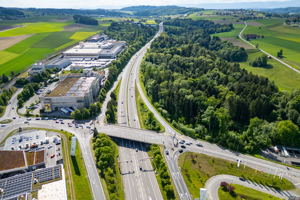 Die Luftaufnahme zeigt eine Autobahnkreuzung in einer grünen Umgebung mit angrenzenden Industriegebäuden. Der Himmel ist klar und die Vegetation saftig grün.