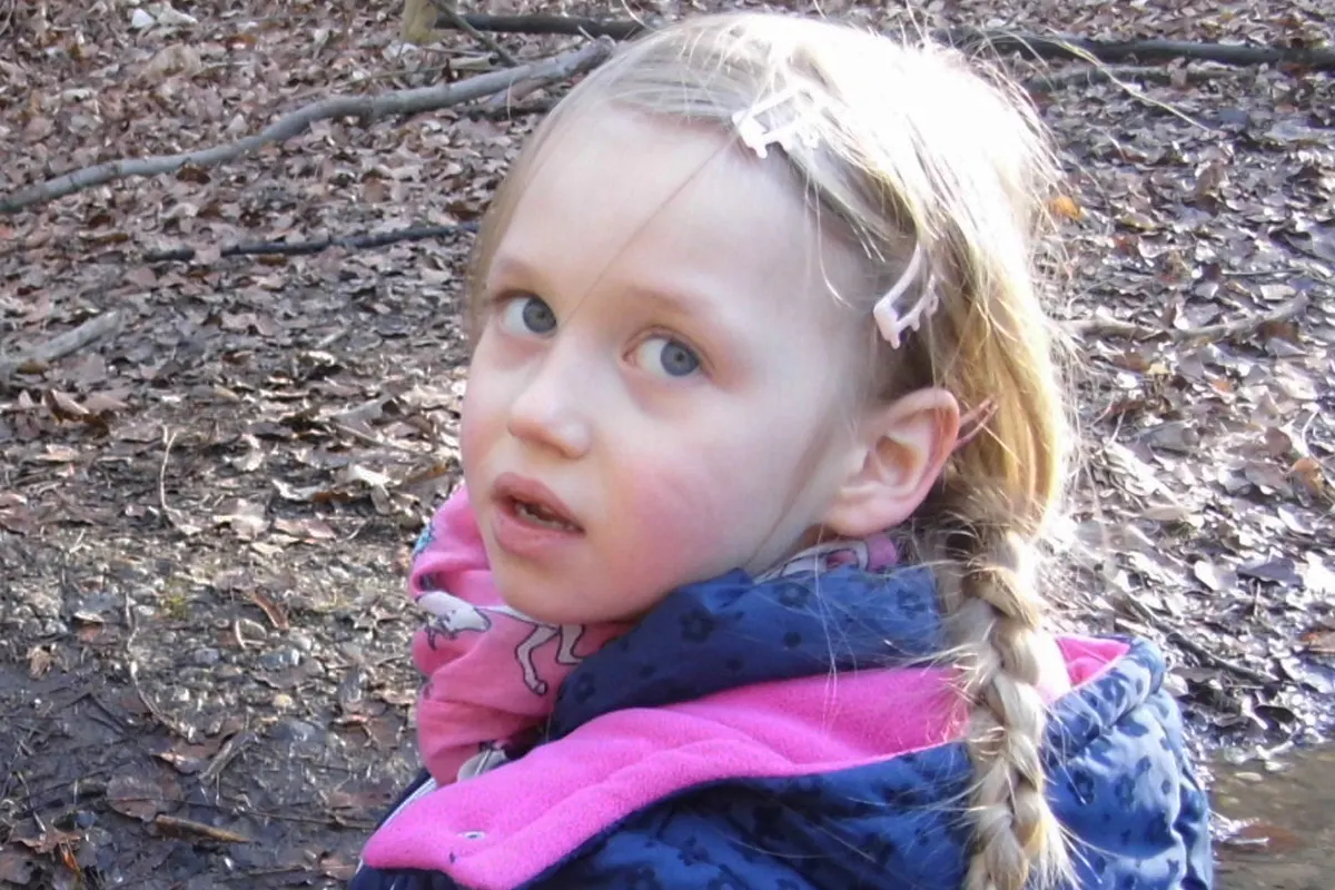 Ein kleines Mädchen mit blonden Zöpfen und pinkem Schal schaut mit ernstem Blick in die Kamera. Sie befindet sich in einem Wald voller Laub.