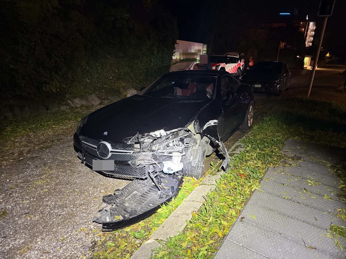 Ein stark beschädigter schwarzer Mercedes mit abgerissener Front steht auf einer unbeleuchteten Straße. Laub bedeckt den Bürgersteig daneben.