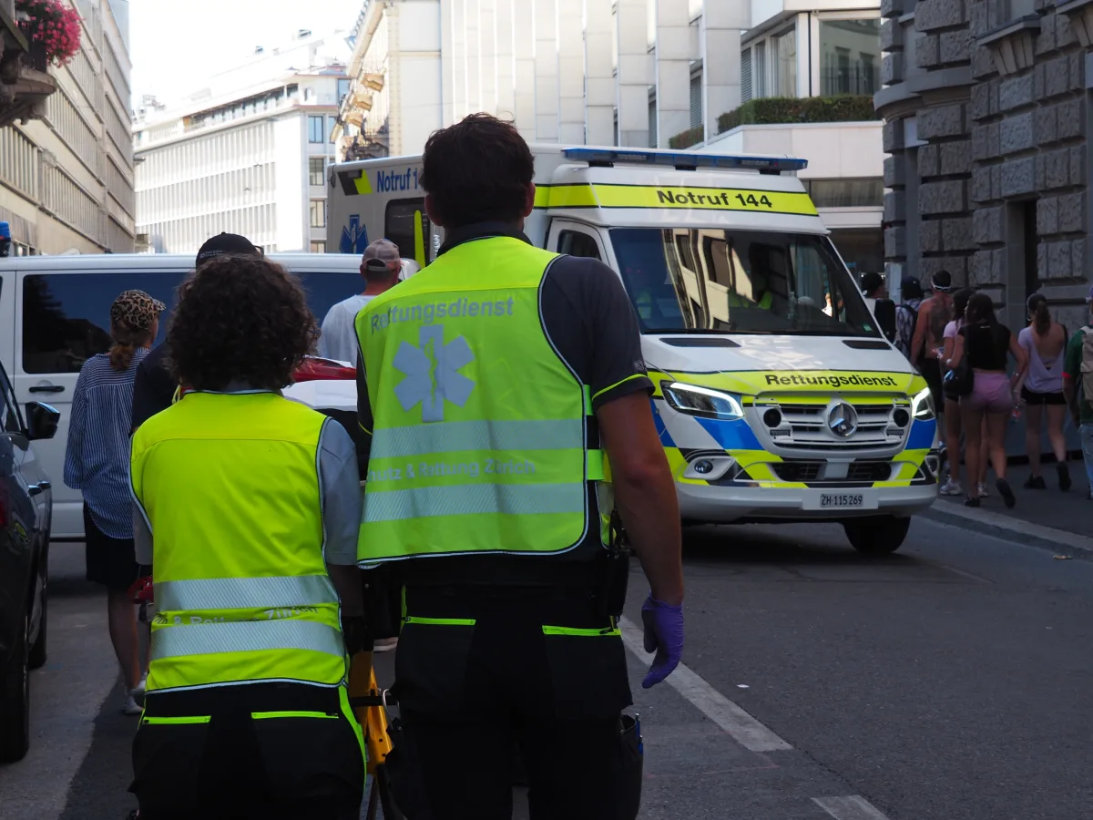 Ein Rettungswagen mit der Aufschrift 'Notruf 144' steht auf einer Straße in Zürich. Zwei Personen in gelben Westen des Rettungsdienstes schauen in Richtung des Wagens, umgeben von Passanten.