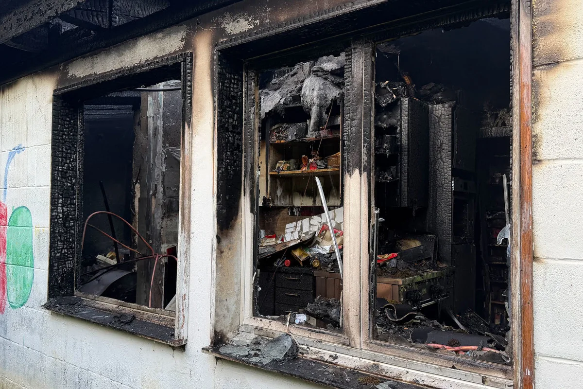 Zwei offene Fenster zeigen die stark verkohlte Innenansicht eines Gebäudes. Ruß- und Brandschäden sind deutlich erkennbar, Möbel sind verbrannt.