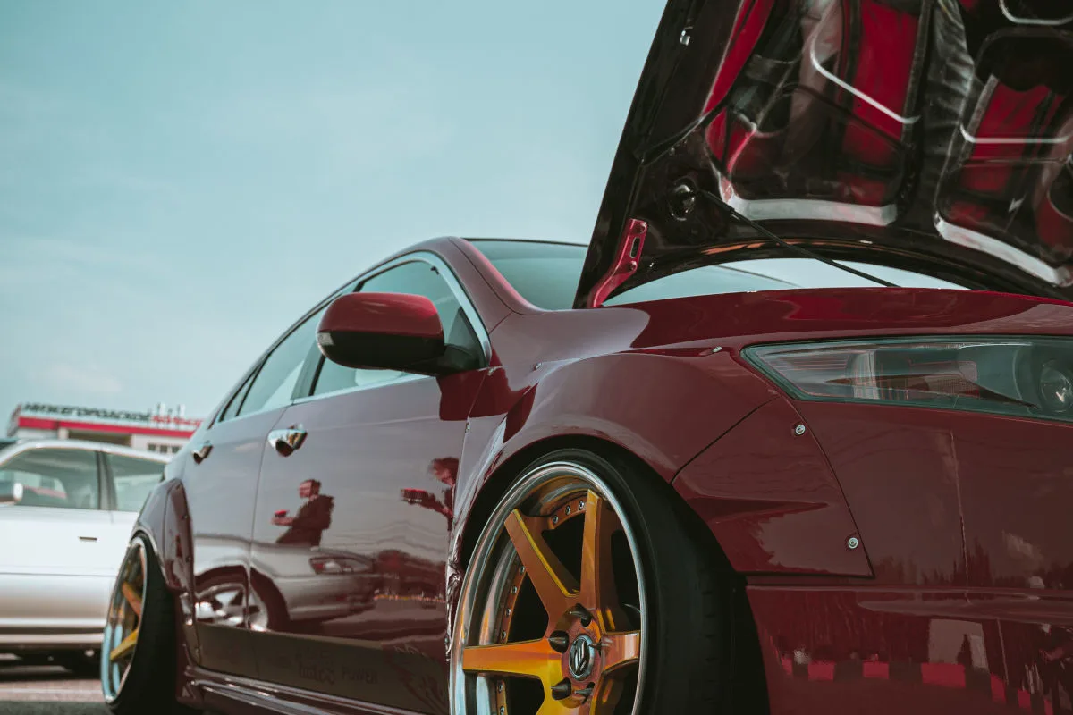 Ein rotes Auto mit geöffneter Motorhaube ist zu sehen. Der Himmel ist klar und im Hintergrund sind weitere Autos erkennbar.