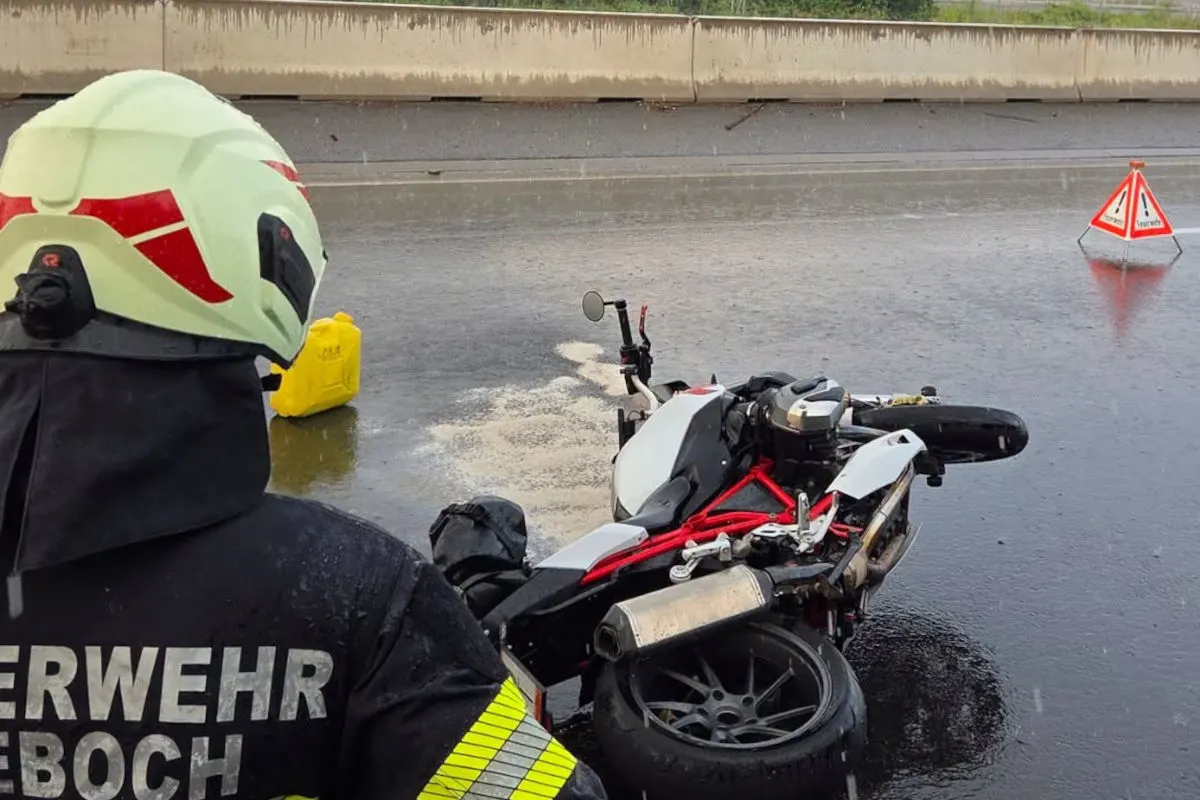 Ein Feuerwehrmann in Schutzkleidung steht vor einem umgestürzten Motorrad auf einer regennassen Autobahn. Das Motorrad liegt quer zur Fahrtrichtung.
