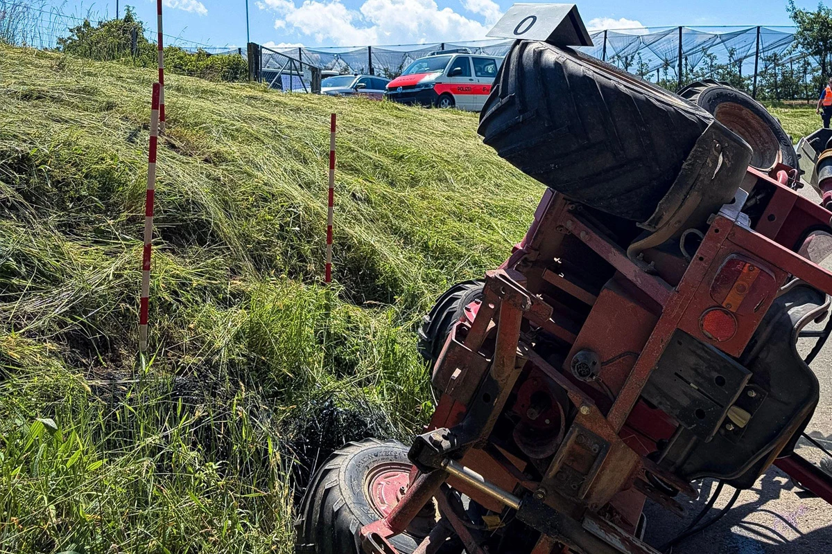 Ein roter Traktor liegt auf der Seite in einem Straßengraben neben einer grasbewachsenen Böschung. Im Hintergrund ist ein Polizeifahrzeug sichtbar.
