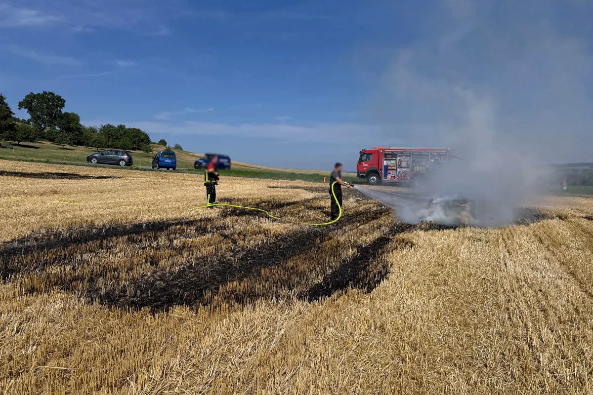 Zwei Feuerwehrleute sind dabei, einen Feldbrand mit einem Wasserschlauch zu löschen, während im Hintergrund ein Feuerwehrfahrzeug steht. Das Feld zeigt sichtbare Brandspuren.