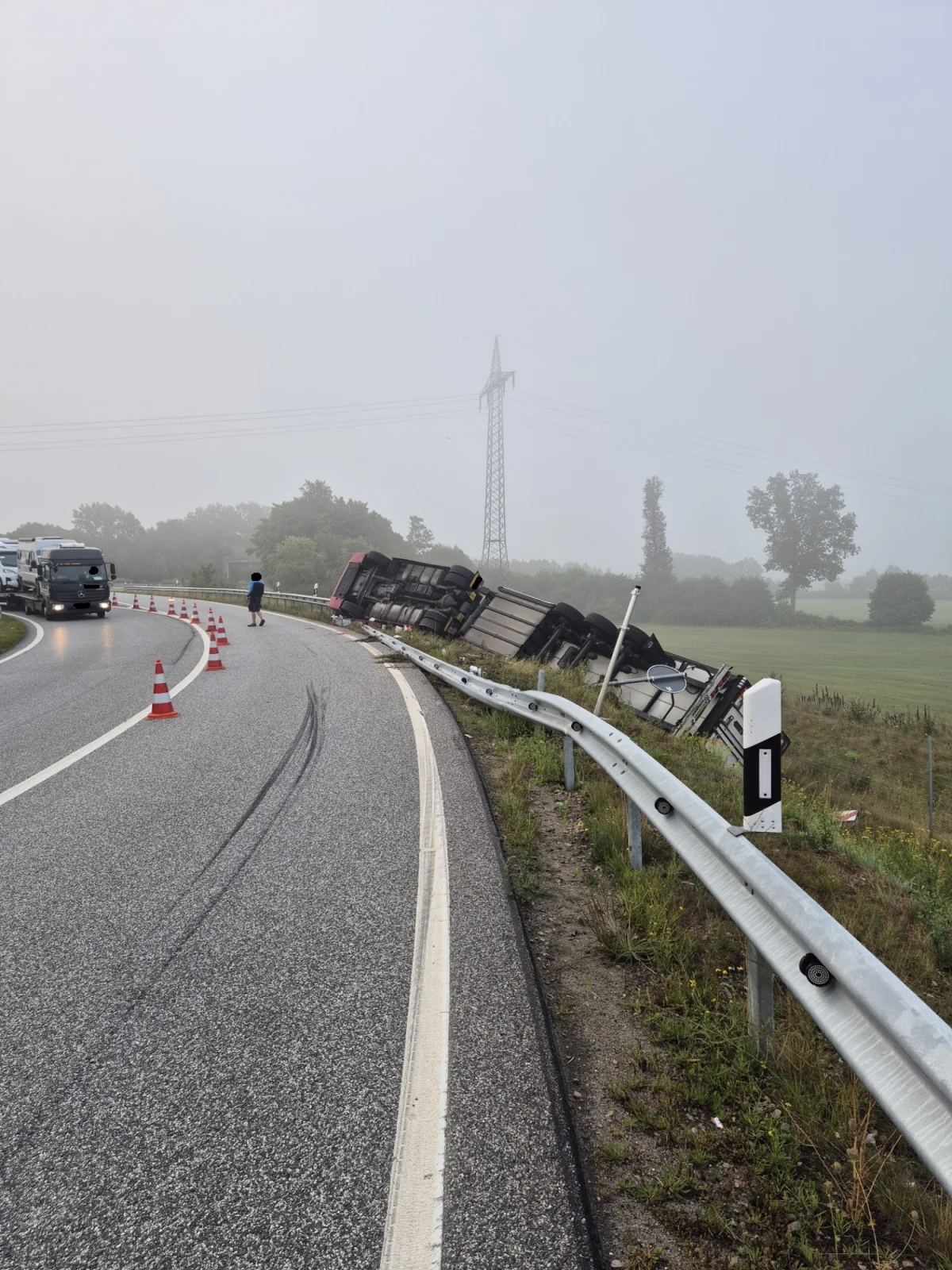 Ein Lastwagen ist von der Straße abgekommen und liegt umgestürzt im Gras. Der Himmel ist neblig und begrenzt die Sicht.