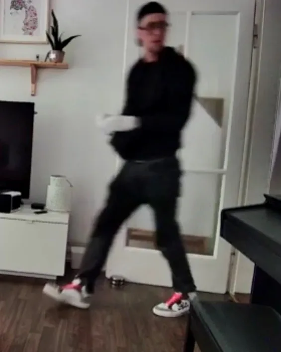 Eine Person in dunkler Kleidung und weißen Handschuhen wird von einer Überwachungskamera in einem Wohnzimmer erfasst. Im Hintergrund sind ein Fernseher, ein Regal mit Deko und ein Schrank sichtbar.