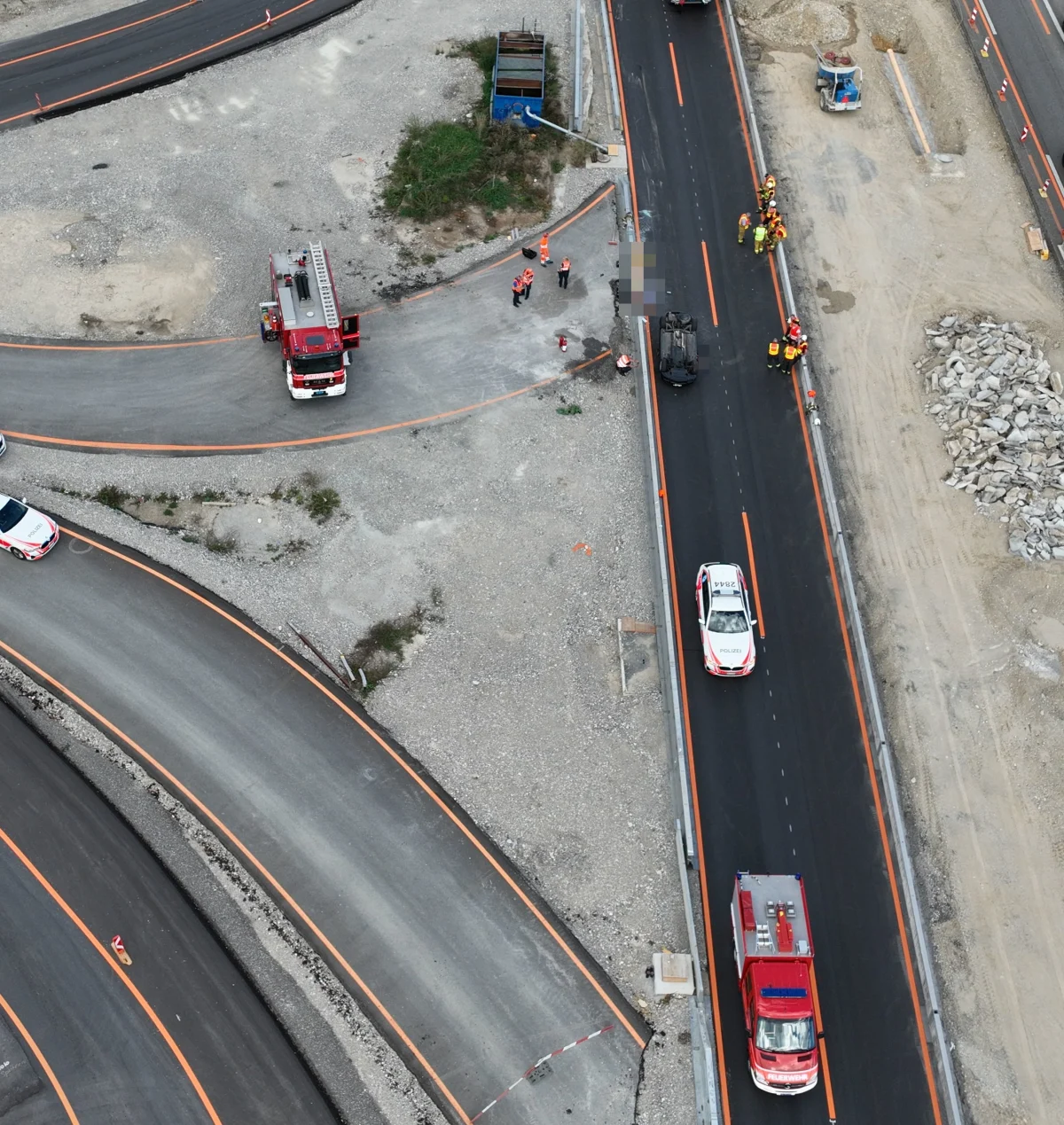Eine Luftaufnahme zeigt ein umgestürztes Fahrzeug auf einer Baustelle. Mehrere Einsatzfahrzeuge und Personen in orangefarbenen Westen sind vor Ort.