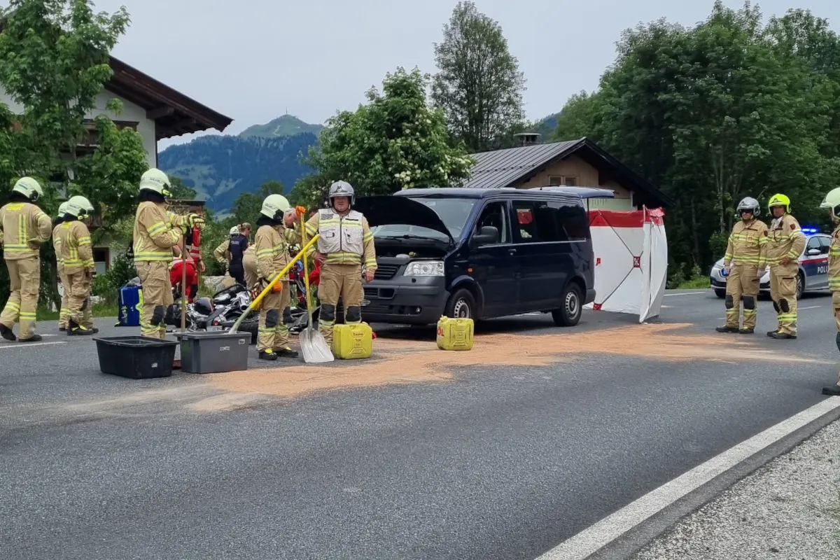 Mehrere Feuerwehrleute in gelber Schutzkleidung stehen um einen blauen Van, dessen Motorhaube offen ist. Sie arbeiten auf einer Landstraße vor einer ländlichen Kulisse mit Bergen im Hintergrund.