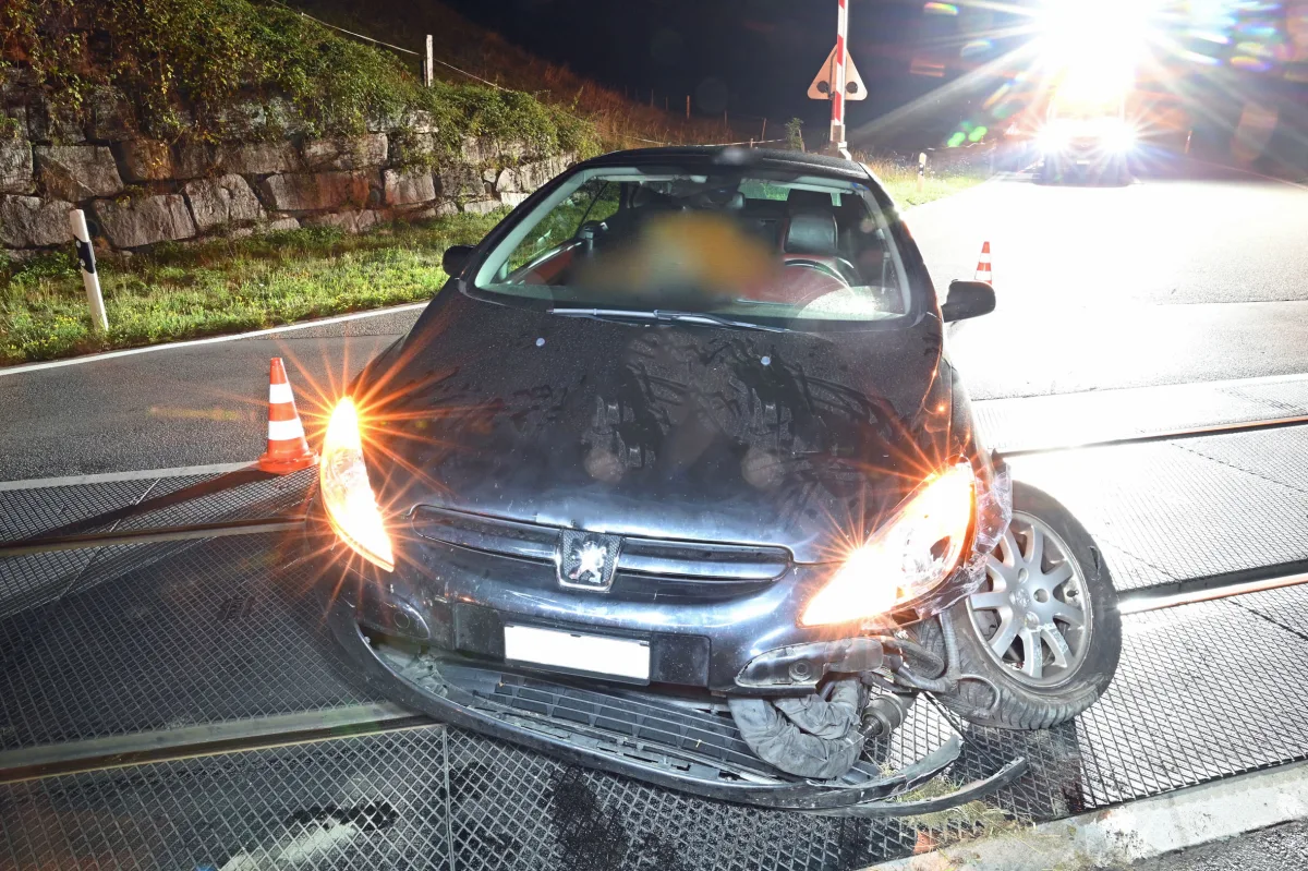 Ein schwarzes Auto steht mit beschädigter Vorderseite auf Bahngleisen. Es ist Nacht, Scheinwerfer und Warnleuchten blenden.