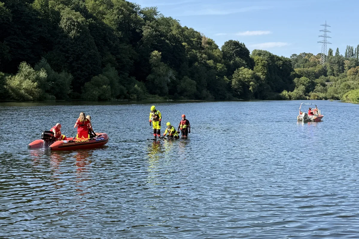 Mehrere Rettungskräfte in leuchtender Schutzkleidung sind auf einem Fluss in einem Rettungsboot und waten im Wasser. Im Hintergrund ist bewaldetes Ufer zu sehen.