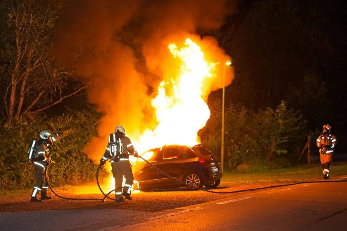 Mehrere Feuerwehrleute in Einsatzkleidung löschen ein stark brennendes Auto auf einer Straße in der Nacht.