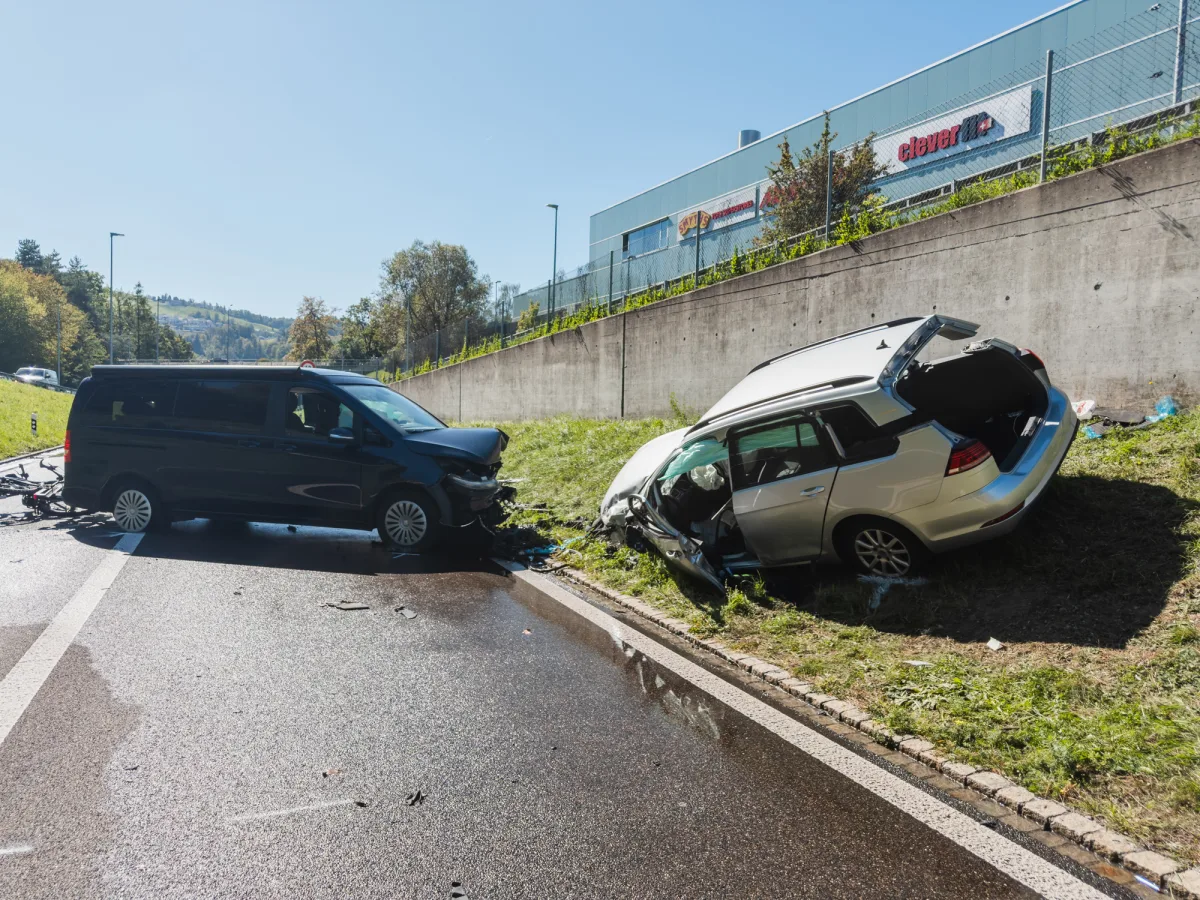 Ein schwarzer Van steht beschädigt auf der Straße, während ein silbernes Auto im Graben liegt. Beide Fahrzeuge haben erhebliche Frontschäden.