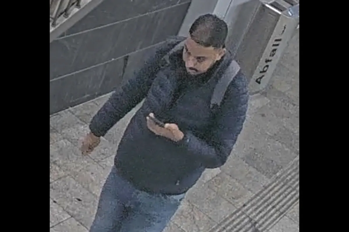 Ein Mann in einer dunklen Jacke und Jeans hält ein Smartphone und läuft durch einen Bahnhofsbereich.