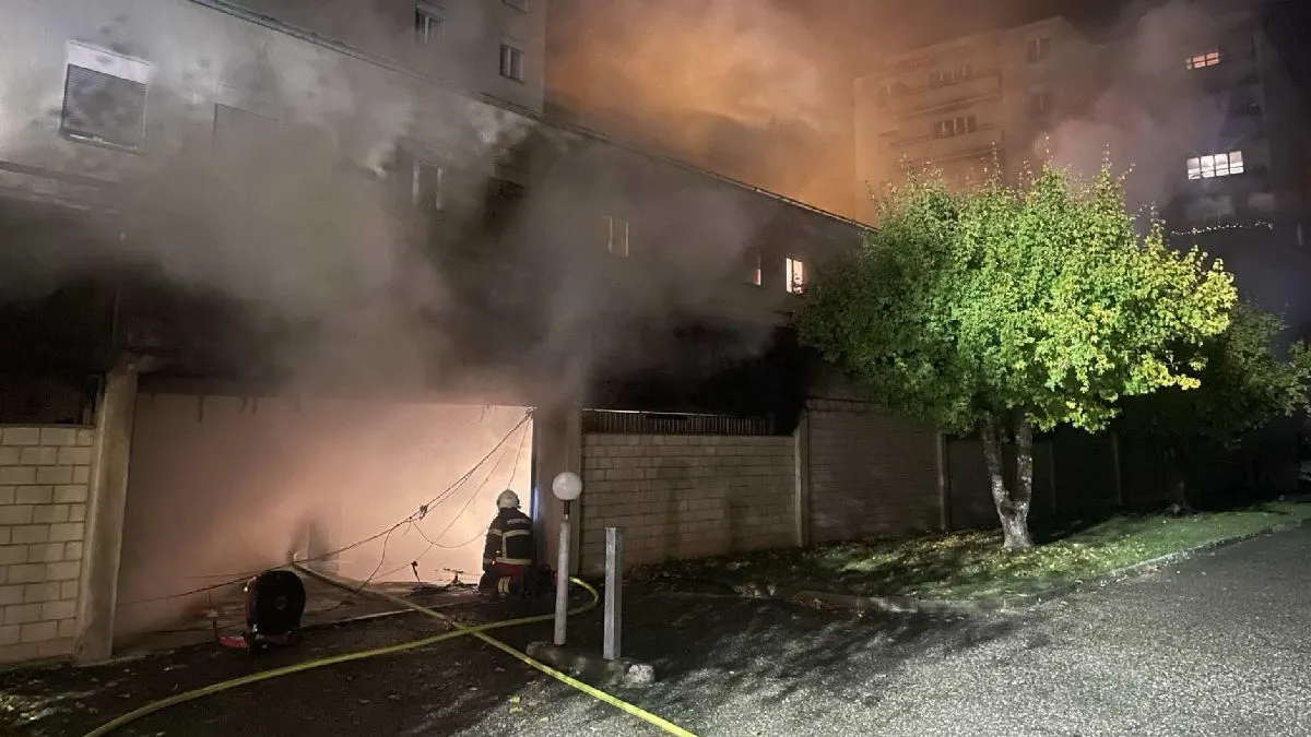 Ein Feuerwehrmann steht vor einer stark rauchenden Garage eines Wohnhauses und arbeitet an der Brandbekämpfung. Dicker Rauch strömt aus der Garage, während der Himmel im Hintergrund durch die Lichter der Umgebung leicht aufgehellt ist.