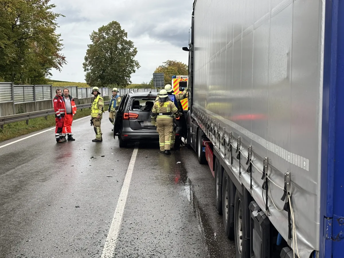 Mehrere Feuerwehrleute und Rettungsdienstmitarbeiter stehen auf einer regennassen Straße neben einem beschädigten Pkw, der neben einem Lkw geparkt ist.