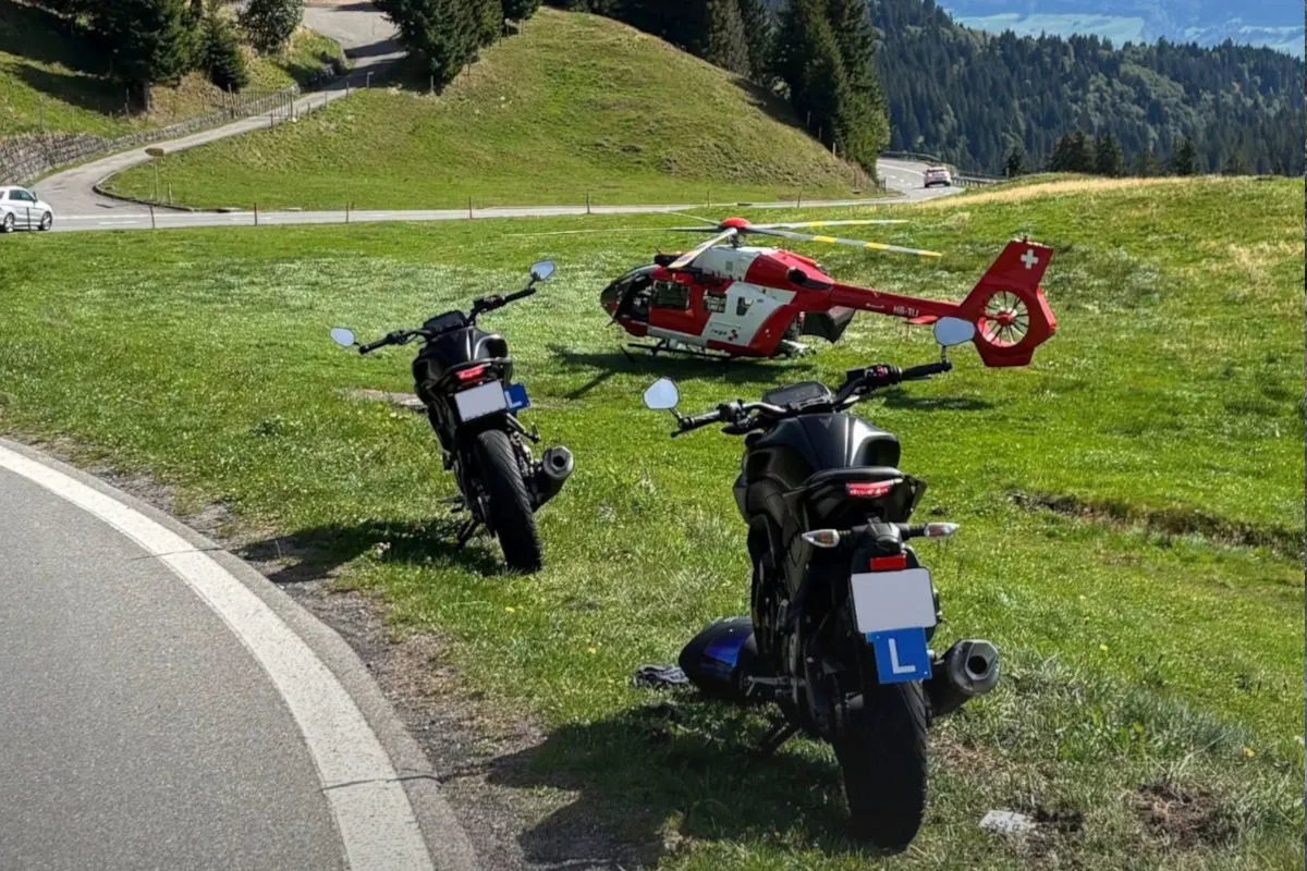 Ein roter Rettungshubschrauber ist auf einer Wiese gelandet. Neben ihm stehen zwei Motorräder. Berge sind im Hintergrund sichtbar.