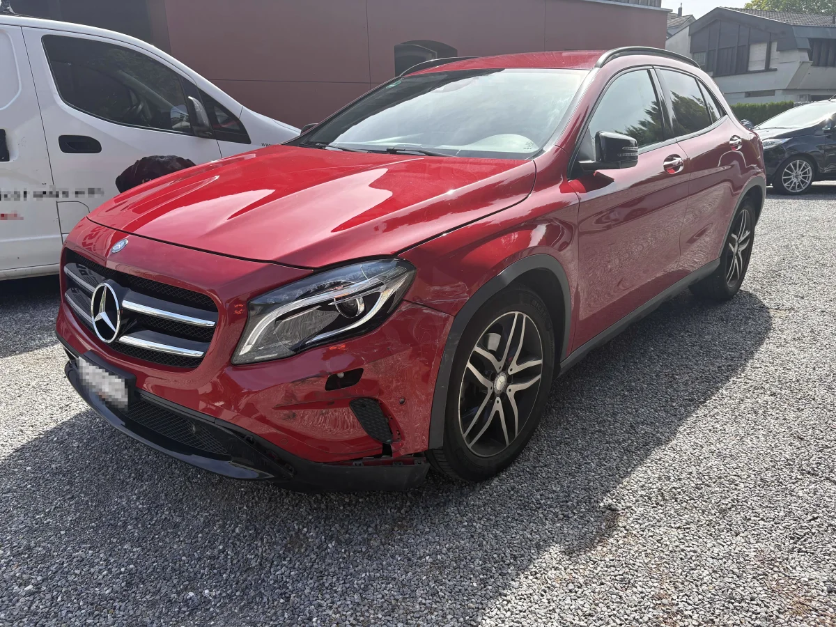 Ein roter Mercedes-Benz zeigt leichte Schäden an der Front und steht auf einem Schotterparkplatz. Im Hintergrund sind andere Fahrzeuge sichtbar.