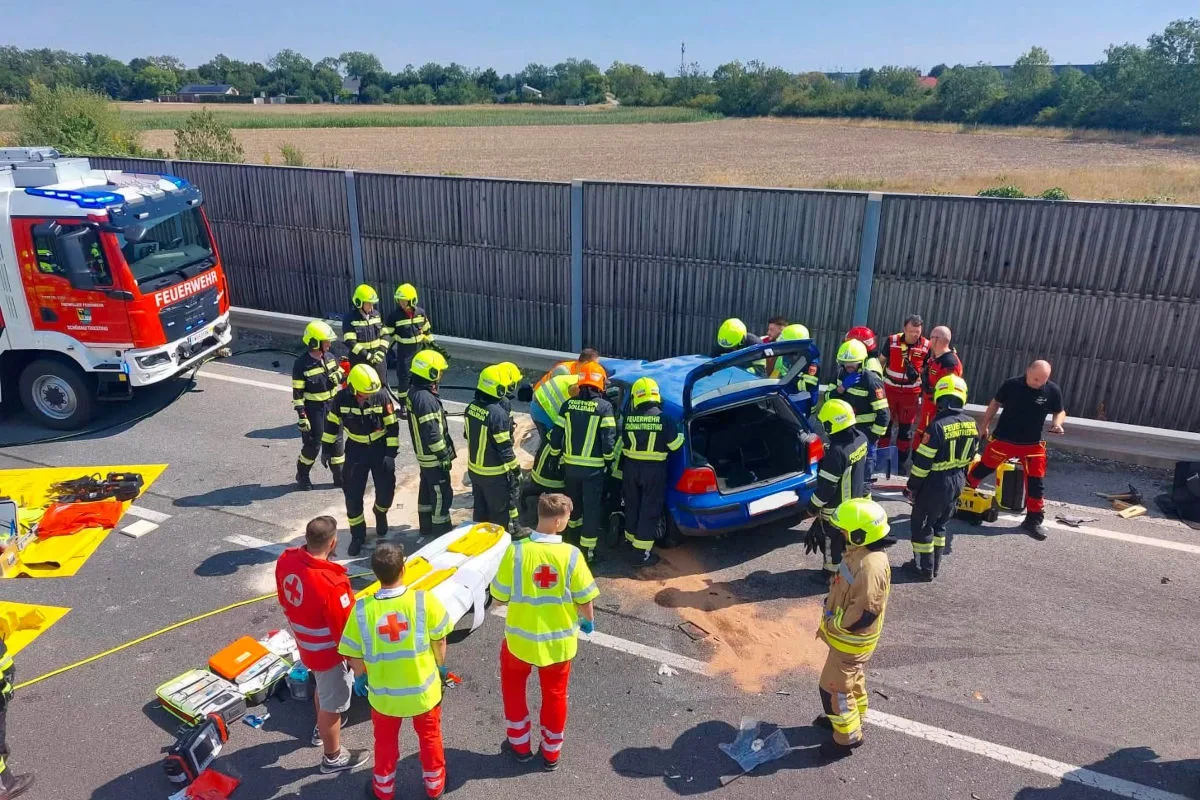 Mehrere Feuerwehrleute und Sanitäter arbeiten an einem blauen Auto auf einer Autobahn. Rettungsgeräte sind um das Fahrzeug verteilt.
