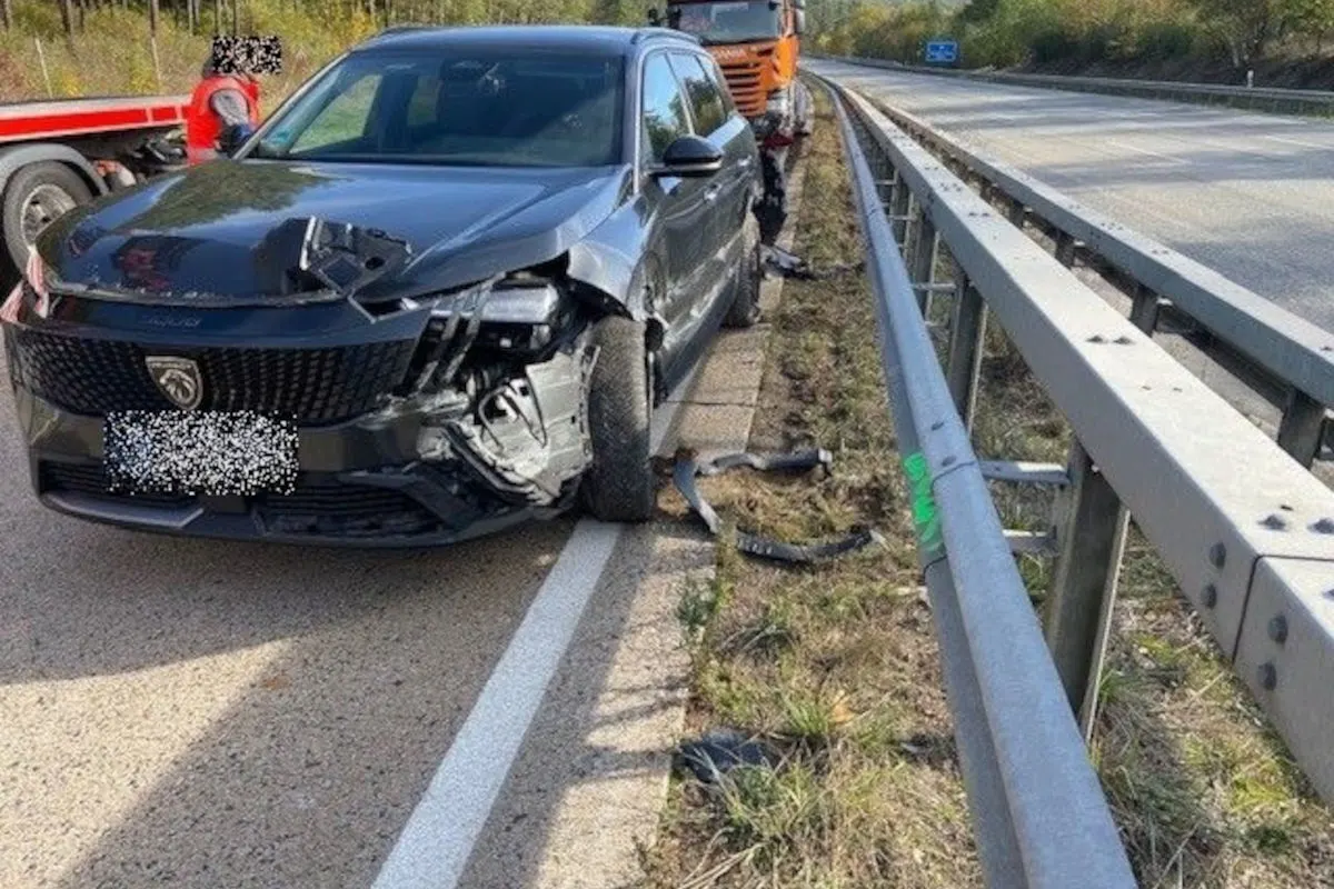 Das Bild zeigt ein stark beschädigtes, dunkles Auto auf einer Autobahn im Freien. Es ist sichtbar mit einem Lkw kollidiert.