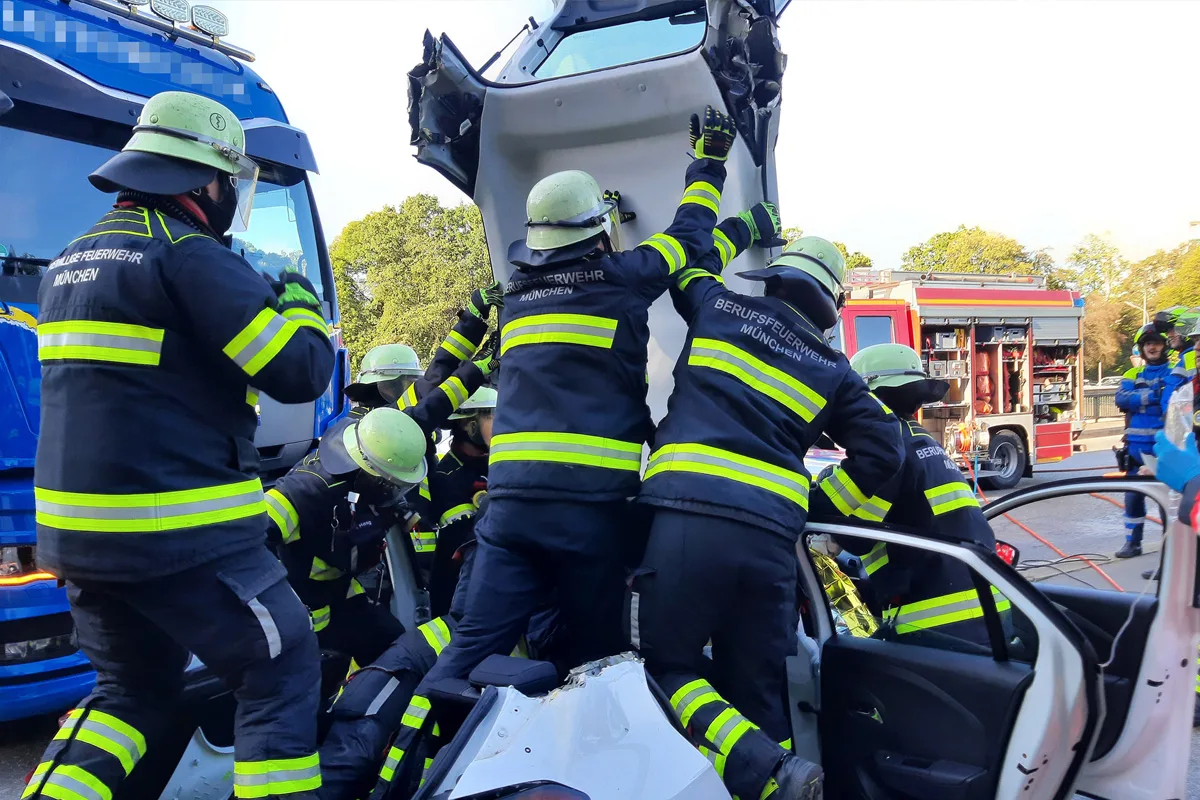 Mehrere Feuerwehrleute, erkennbar an ihrer Uniform, arbeiten an einem beschädigten Fahrzeug auf einer Straße.