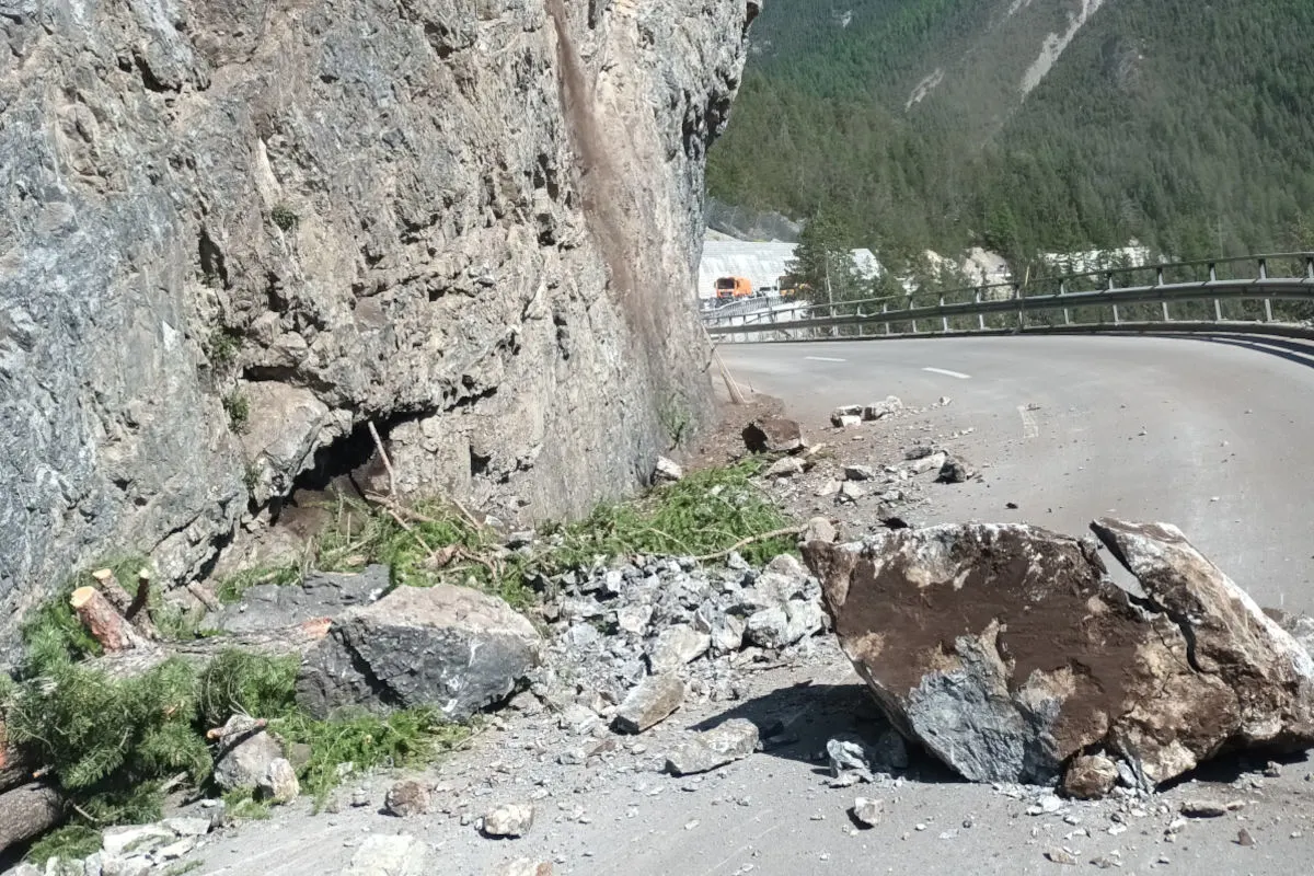 Ein Bereich einer Bergstraße ist durch herabgestürztes Geröll und größere Felsbrocken blockiert. Rechts von der Straße ist eine Leitplanke sichtbar, im Hintergrund sind bergige Landschaften und ein Fahrzeug zu sehen.