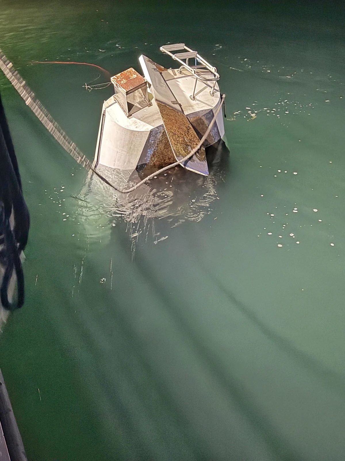 Ein stark geneigtes, teilweise untergetauchtes Boot mit Metallrahmen und verrosteten Teilen schwimmt auf grünlichem Wasser. Ein Seil ist mit dem Boot verbunden.
