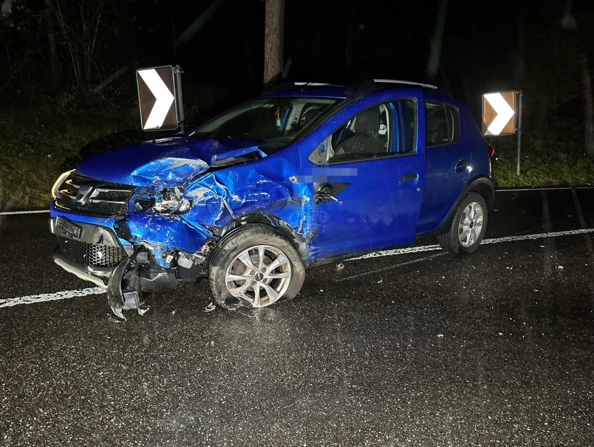 Ein blaues Auto mit schweren Frontschäden steht auf einer nassen Straße. Die Umgebung ist dunkel, die Beleuchtung begrenzt.