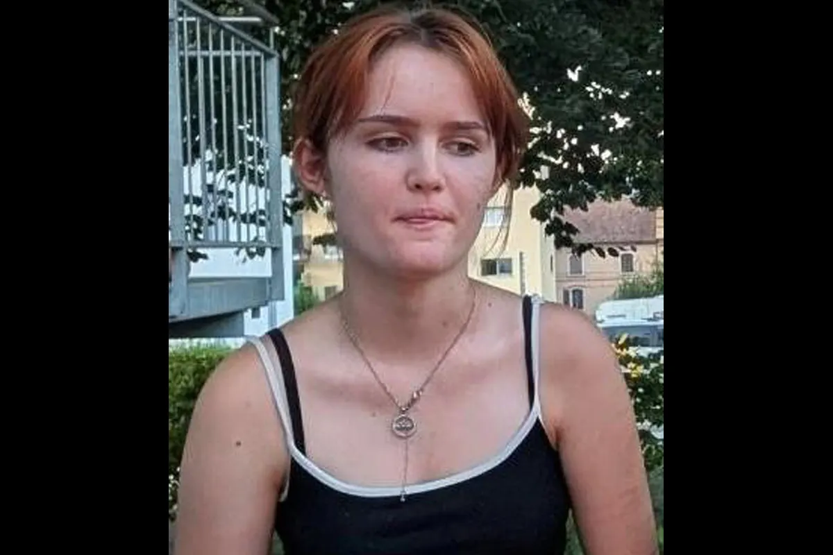 Das Bild zeigt eine junge Frau mit rötlichen Haaren, die ein dunkles Top trägt. Im Hintergrund sind Gebäude und Bäume sichtbar.