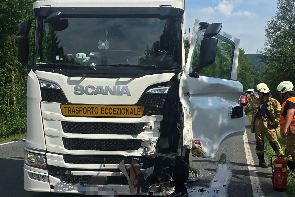 Ein weißer Scania-Lkw, stark beschädigt an der Front, steht auf einer Landstraße. Feuerwehrleute in Schutzkleidung sind nahebei.