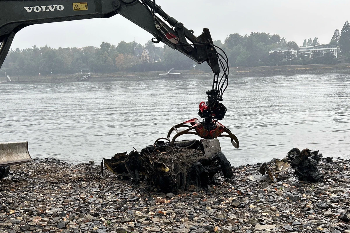 Ein Bagger mit Greifarm hebt ein rostiges Autowrack am kiesigen Ufer eines Flusses. Im Hintergrund sind Bäume und Gebäude am anderen Ufer zu sehen.