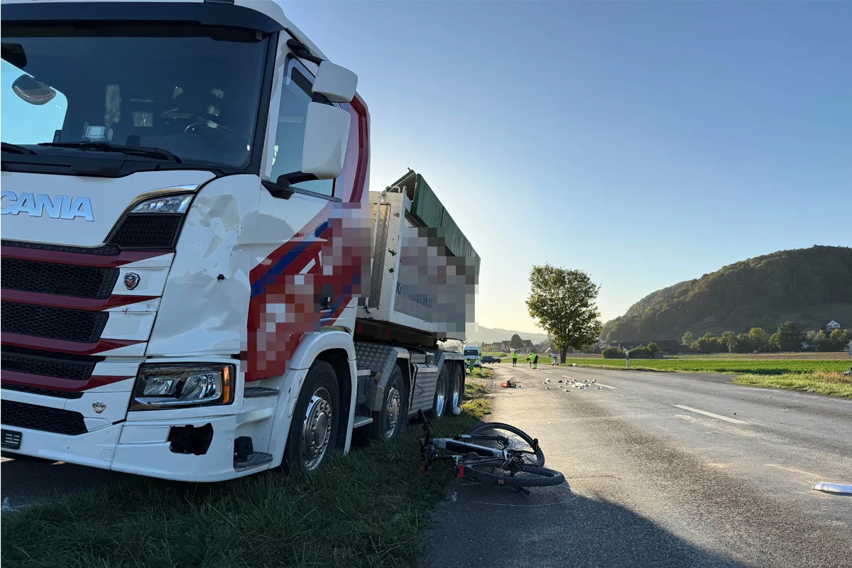 Ein weißer Lastwagen mit Schäden an der Vorderseite steht auf einer Landstraße neben einem umgestürzten Fahrrad. Im Hintergrund ist eine ländliche Umgebung sichtbar.