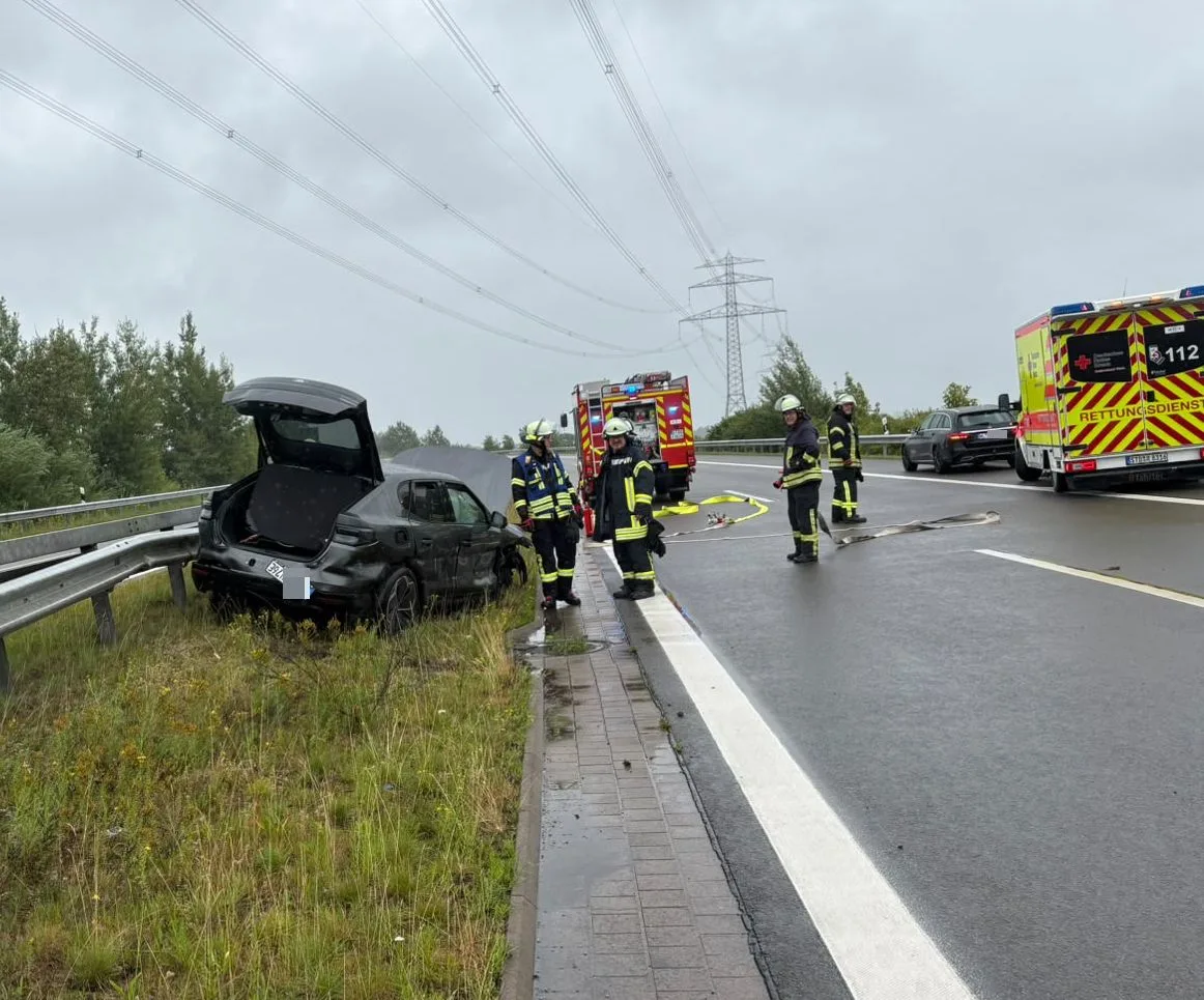 Ein Auto mit geöffnetem Kofferraum steht beschädigt an der Leitplanke. Feuerwehrleute arbeiten vor Ort, während ein Rettungswagen in der Nähe parkt.