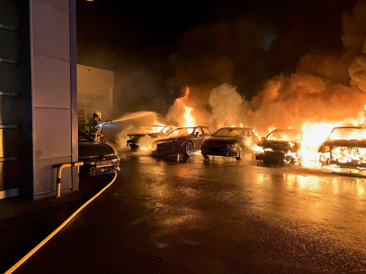 Ein Feuerwehrmann bekämpft bei Nacht die Flammen an mehreren brennenden Fahrzeugen auf einem Parkplatz. Das Feuer erzeugt dichten Rauch und intensive Hitze.