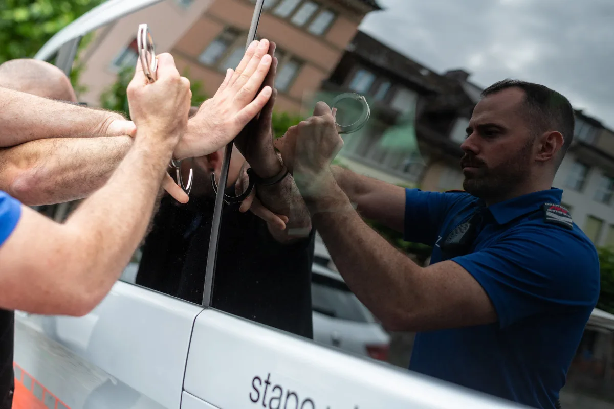 Ein Polizist legt einer Person Handschellen an, während deren Hände gegen ein Autofenster gedrückt sind. Die Szene spiegelt sich im Fenster, ein Streifenwagen ist sichtbar.