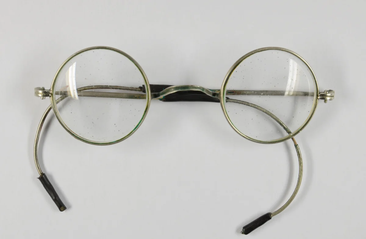 Das Bild zeigt eine alte Brille mit runden Gläsern und abgenutzten Bügeln auf einem weißen Hintergrund.