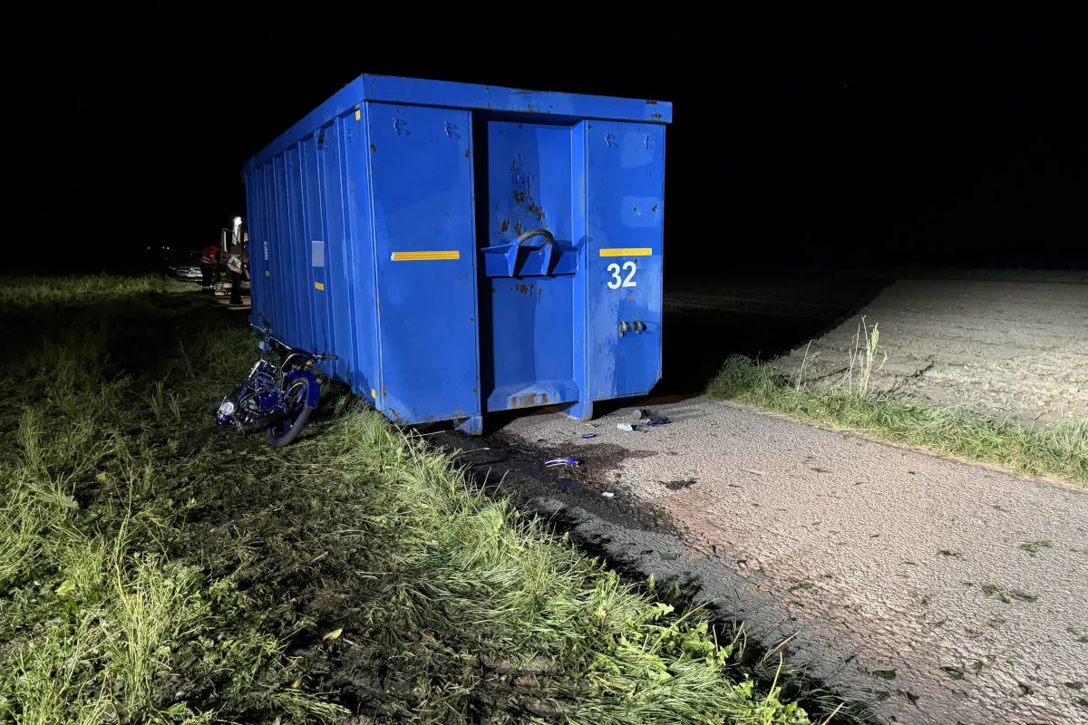 Bei Nacht liegt ein blaues Motorrad beschädigt neben einem großen blauen Container auf einer ländlichen Straße. Der Container steht auf einer Grasfläche, die Straße wirkt abgelegen.