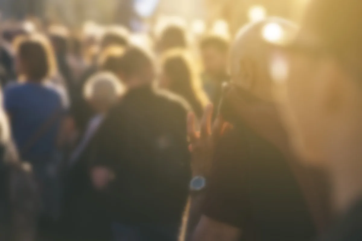 Eine unscharfe Menschenmenge bei Tageslicht, eine Person telefoniert. Der Fokus liegt auf einem Mann im Vordergrund.