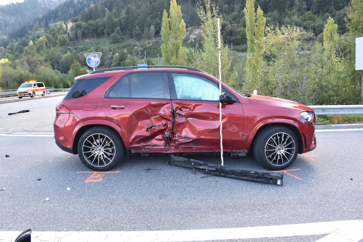 Ein roter SUV mit schweren Schäden an der Fahrerseite steht auf einer Landstraße. Im Hintergrund sind ein Notfallfahrzeug und Bäume sichtbar.
