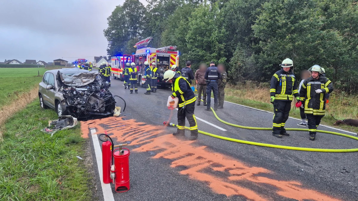 Ein stark beschädigtes Auto am Straßenrand, umgeben von Feuerwehrleuten, die die Unfallstelle sichern. Im Hintergrund sind weitere Einsatzfahrzeuge zu sehen.