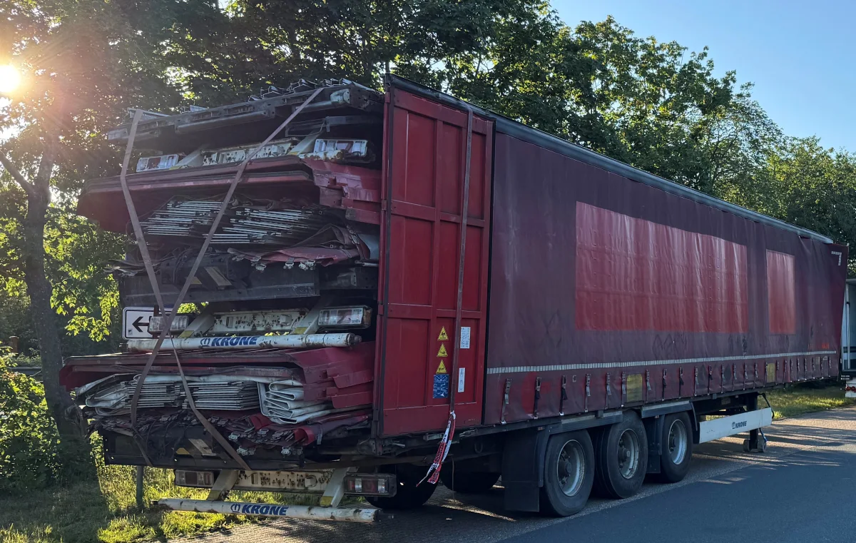 Ein roter Lkw-Anhänger steht am Straßenrand und transportiert gestapelte, stark beschädigte Autowracks. Die Umgebung ist grün und bewaldet, mit Sonnenschein im Hintergrund.