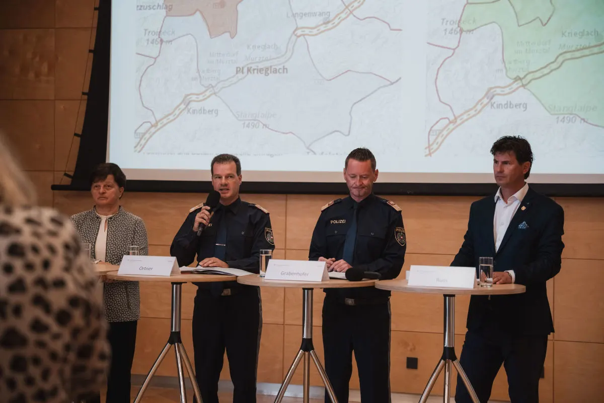 Vier Personen stehen oder sitzen an Rednerpulten, zwei davon in Polizeiuniformen, vor einer projizierten Karte.