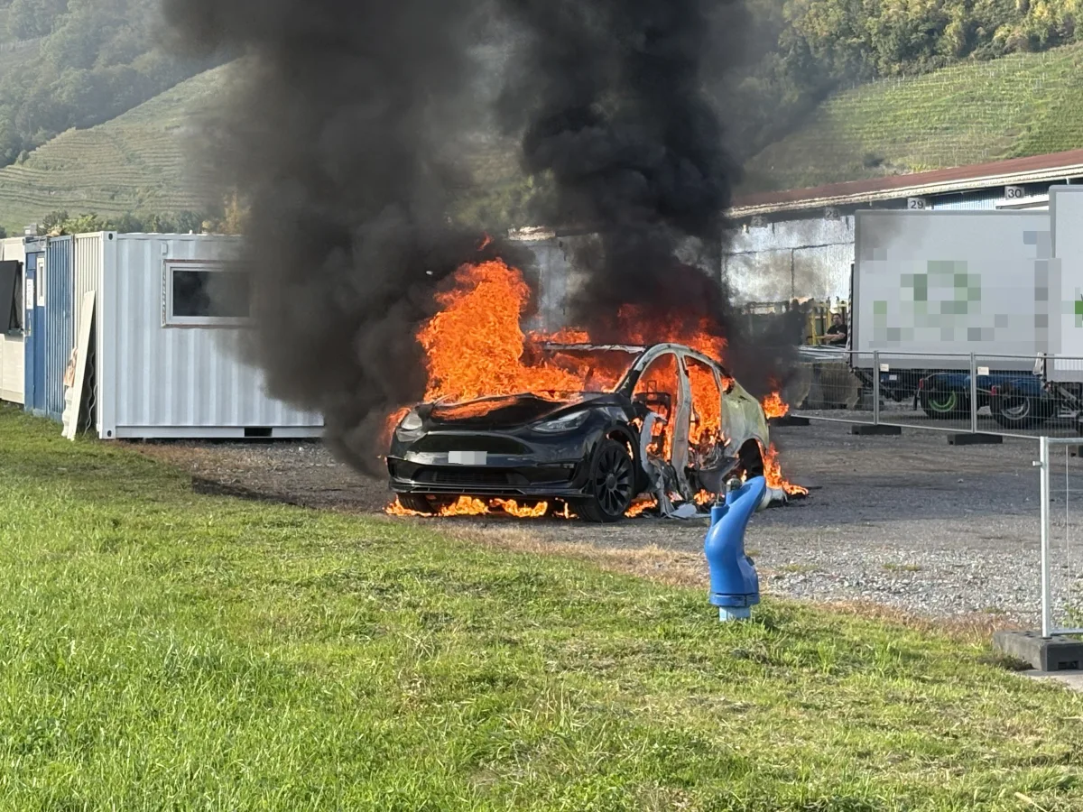 Ein Auto steht lichterloh in Flammen, große Rauchwolken steigen auf, neben ihm sind Container und grüne Wiesen zu sehen.