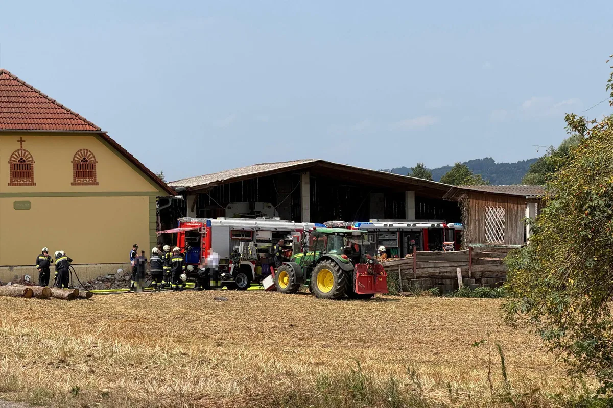 Feuerwehrleute und Einsatzfahrzeuge stehen vor einem landwirtschaftlichen Gebäude auf einem abgeernteten Feld. Ein Traktor ist ebenfalls sichtbar.