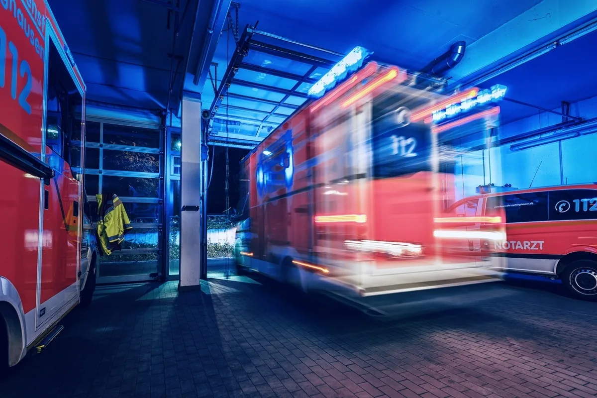 In einer hell erleuchteten Garage fährt ein Rettungsfahrzeug mit eingeschaltetem Blaulicht hinaus. Rechts ist ein weiteres Fahrzeug mit der Aufschrift 'Notarzt' zu sehen.
