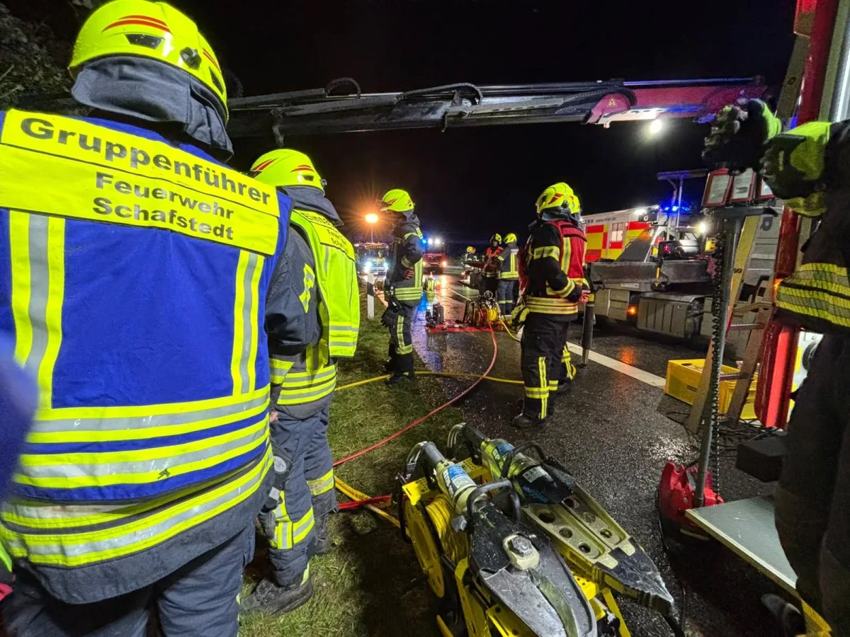 Mehrere Feuerwehrkräfte in Schutzbekleidung arbeiten bei Nacht auf einer nassen Straße an einer Unfallstelle, daneben Rettungsfahrzeuge.